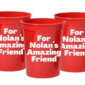 Puede incluir: Tres vasos de pl&aacute;stico rojos con texto blanco que dice "Para el incre&iacute;ble amigo de Nolan".