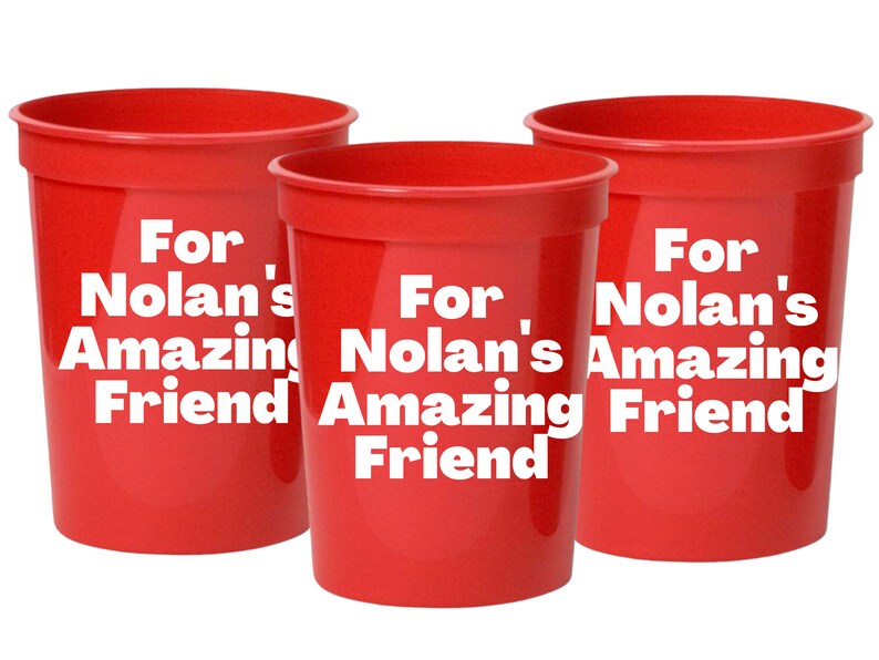 Puede incluir: Tres vasos de pl&aacute;stico rojos con texto blanco que dice "Para el incre&iacute;ble amigo de Nolan".