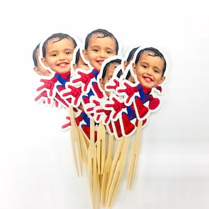 Puede incluir: Un conjunto de seis adornos para tartas de Spiderman, rojos y azules, con una foto de la cara de un ni&ntilde;o en cada uno. Los adornos est&aacute;n unidos a brochetas de madera.