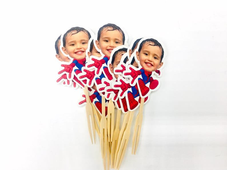 Puede incluir: Un conjunto de seis adornos para tartas de Spiderman, rojos y azules, con una foto de la cara de un ni&ntilde;o en cada uno. Los adornos est&aacute;n unidos a brochetas de madera.