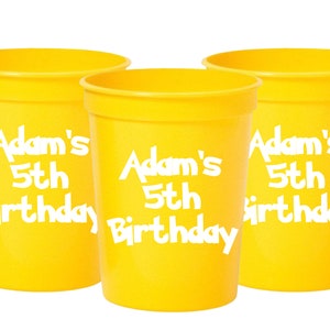 Pu&ograve; includere: Tre bicchieri di plastica gialli con testo bianco che dice "Adam's 5th Birthday".