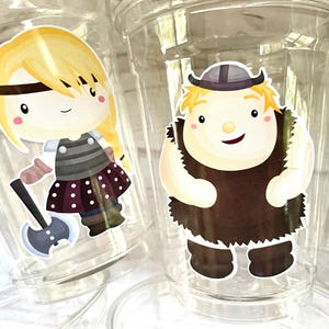 May include: Two clear plastic cups with colorful cartoon stickers. One sticker features a blonde girl with a braid, wearing a striped shirt and a polka dot skirt, holding an axe. The other sticker features a blonde boy with a beard, wearing a brown tunic and a horned helmet.