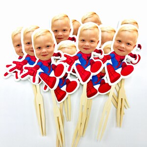 Puede incluir: Un conjunto de 10 adornos para cupcakes de Spiderman rojos y azules con la cara de un ni&ntilde;o en cada uno. Los adornos est&aacute;n en pinchos de madera con un tenedor en el extremo.