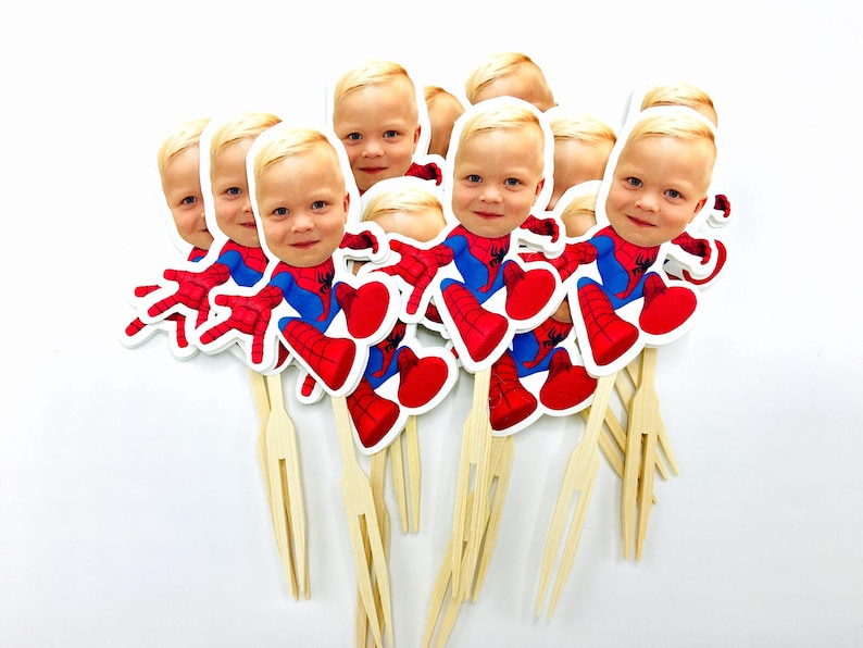 Puede incluir: Un conjunto de 10 adornos para cupcakes de Spiderman rojos y azules con la cara de un ni&ntilde;o en cada uno. Los adornos est&aacute;n en pinchos de madera con un tenedor en el extremo.