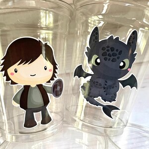 May include: Two clear plastic cups with cartoon character designs. One cup features a black dragon with a white outline and the other cup features a cartoon boy with brown hair holding a shield.