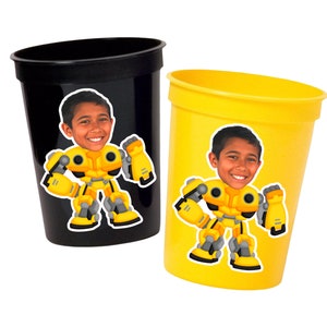 May include: Two yellow plastic cups with a black rim. Each cup has a sticker of a cartoon robot with a person's face on it. The robot is yellow with black and gray details.