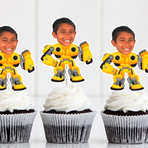 May include: Three yellow and gray robot cupcake toppers with a person's face on the head. The toppers are on top of chocolate cupcakes with white frosting.