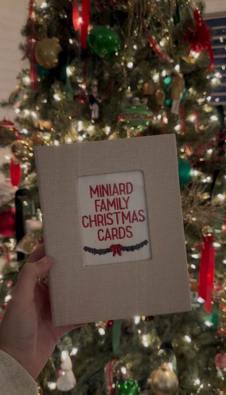 May include: A rectangular beige fabric-covered photo album with a central white window containing the red text MINIARD FAMILY CHRISTMAS CARDS above a small green garland graphic with a red bow, designed for storing holiday greeting cards.