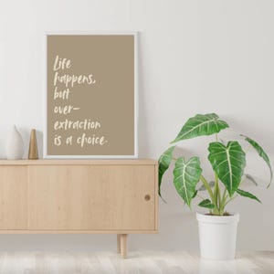 May include: A framed print with a beige background and white text that reads "Life happens, but over-extraction is a choice." The print is hanging on a white wall above a light wood cabinet with a potted green plant in front of it.