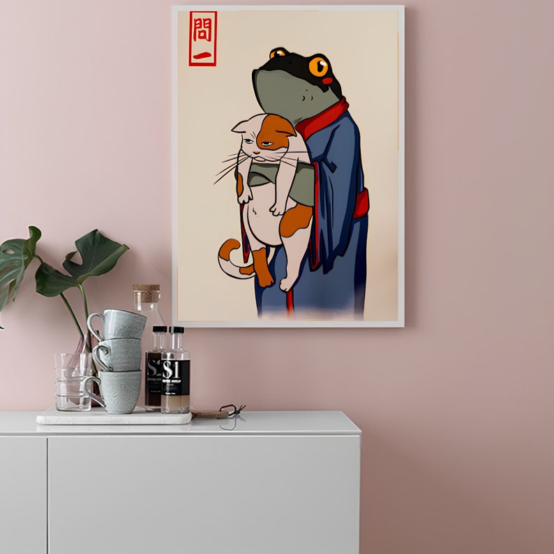 Op de afbeelding: Een ingelijste kunstdruk met een cartoonkikker in een blauwe jurk die een kat vasthoudt. De kat is wit en oranje. De kikker heeft een zwarte kop en gele ogen. Japanse tekst in de linkerbovenhoek.