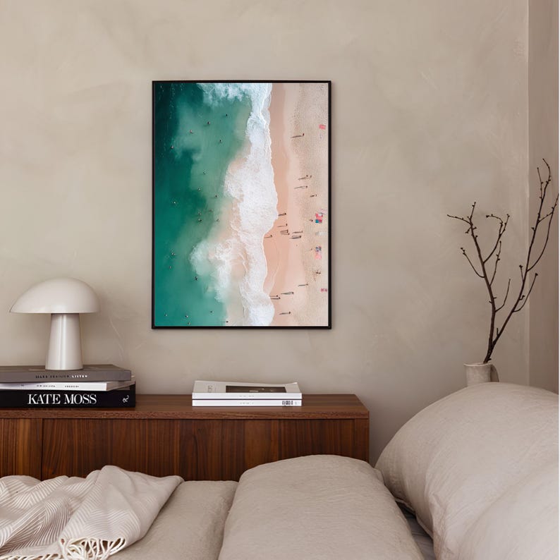 May include: An aerial photograph of a beach with turquoise water and white waves crashing on the shore. The sand is a light pink color and there are many people swimming and sunbathing.