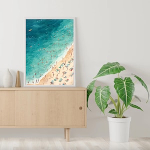 May include: A framed watercolor painting of a beach scene with people swimming and sunbathing. The painting is in a white frame and is hanging on a wall.