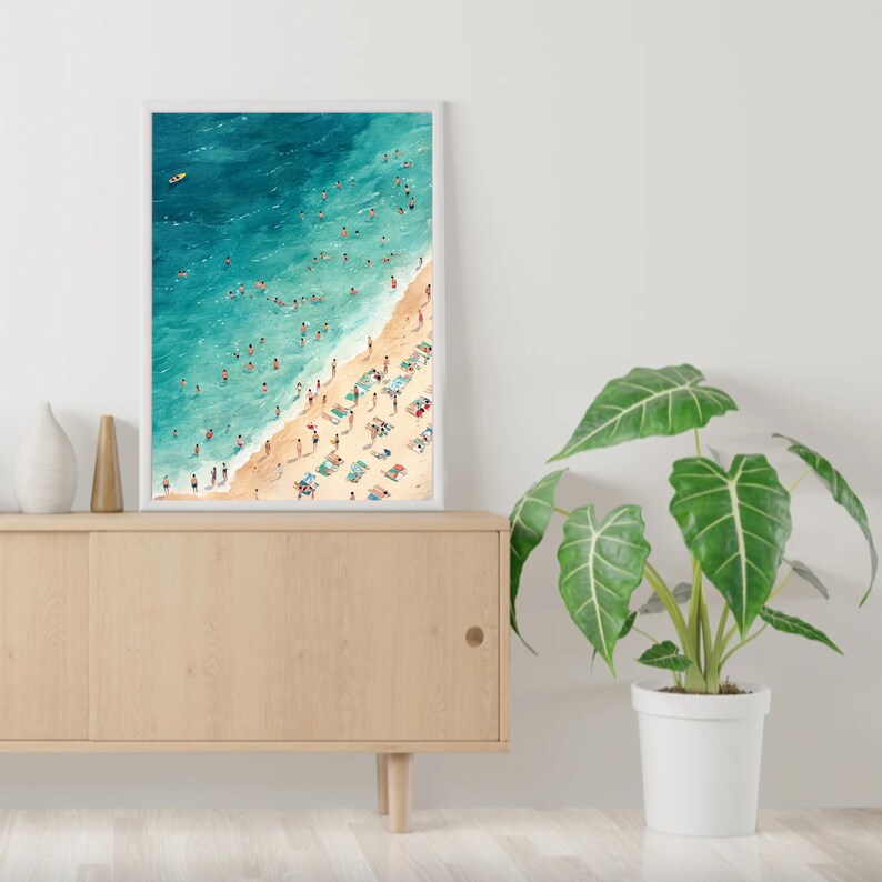 May include: A framed watercolor painting of a beach scene with people swimming and sunbathing. The painting is in a white frame and is hanging on a wall.