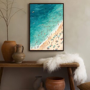 May include: An aerial view watercolor painting of a beach scene with people swimming and sunbathing on the sand. The water is a vibrant blue and the sand is a light beige. The painting is framed in a black frame.