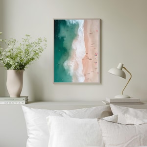 May include: An aerial photograph of a beach with turquoise water and white waves crashing on the shore. The sand is light pink and there are many people on the beach.