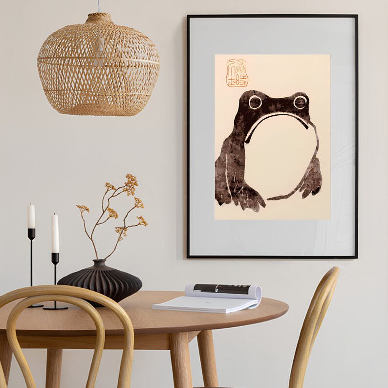 Op de afbeelding: Een ingelijste kunstprint met een gestileerde kikkerillustratie in bruin- en cr&egrave;metinten. Het kunstwerk heeft een minimalistische esthetiek met een Japans ge&iuml;nspireerd ontwerp. De print wordt gepresenteerd in een zwarte lijst.