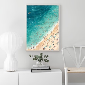 May include: A watercolor painting of a beach scene with people swimming and sunbathing. The water is a vibrant blue and the sand is a light beige. The painting is framed in a white frame.