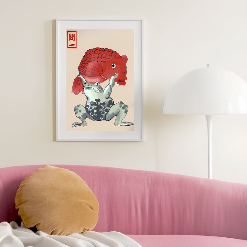 Op de afbeelding: Ingelijste kunstprint met een grillige illustratie van een kikker die een grote rode vis boven zich houdt. De kikker is groen met donkere vlekken en de vis heeft een gedetailleerd, gestructureerd uiterlijk. Het kunstwerk staat tegen een cr&egrave;mekleurige achtergrond, met Japanse tekst.