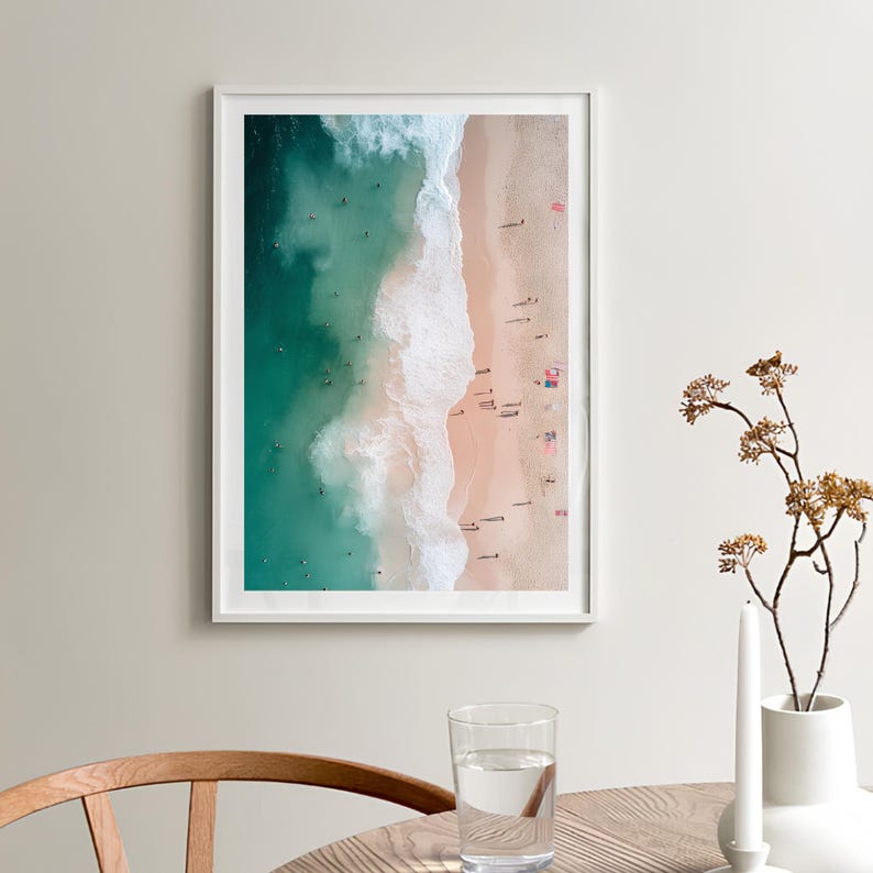 May include: An aerial photograph of a beach with turquoise water and white waves crashing on the shore. The sand is light pink and there are many people swimming and sunbathing.