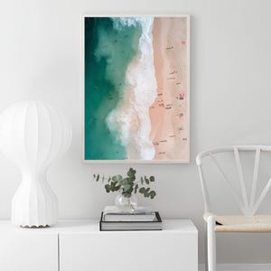 May include: An aerial photograph of a beach with turquoise water and white waves crashing on the shore. The sand is light pink and there are many people on the beach.