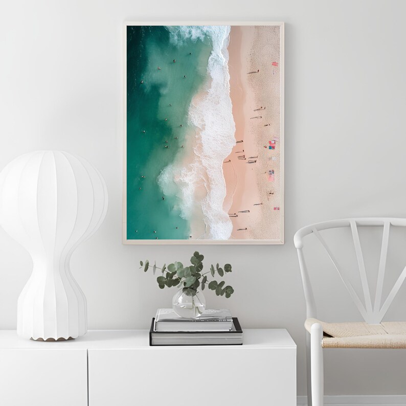 May include: An aerial photograph of a beach with turquoise water and white waves crashing on the shore. The sand is light pink and there are many people on the beach.