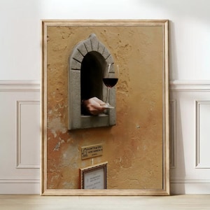 May include: A framed print depicts a hand holding a glass of red wine through a stone window. The window is set in a textured, ochre-colored wall. A small sign below the window reads "Ricettina del Vino Wine Window".