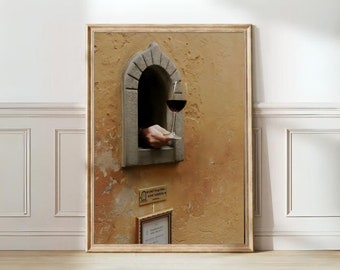 May include: A framed print depicts a hand holding a glass of red wine through a stone window. The window is set in a textured, ochre-colored wall. A small sign below the window reads "Ricettina del Vino Wine Window".