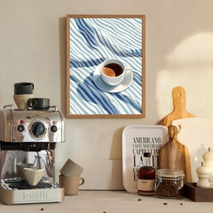 May include: A framed illustration of a cup of coffee on a blue and white striped tablecloth. The cup is white with a brown rim and a brown handle. The coffee is dark brown. The tablecloth is folded over itself and the stripes are visible. The frame is brown wood.