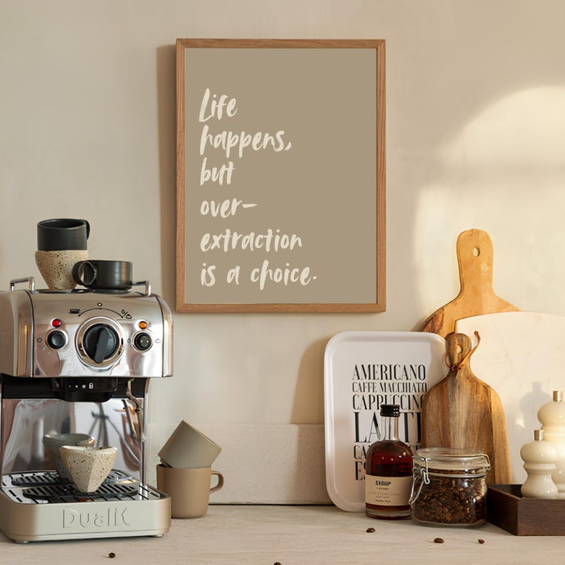 May include: A framed print with the text "Life happens, but over-extraction is a choice."  The print is on a beige background with white text. The print is hanging on a wall above a coffee maker and other coffee-related items.