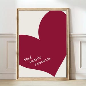 Op de afbeelding: Ingelijste kunstprint met een groot bordeauxrood hart op een gebroken witte achtergrond. De tekst in cursief in het hart luidt "Qué suerte tenemos". De lijst heeft een lichte houtkleur.
