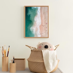 May include: A framed photograph of an aerial view of a beach with turquoise water and white waves crashing on the shore. The sand is light pink and there are people swimming and sunbathing. A stuffed koala toy is sitting in a woven basket.