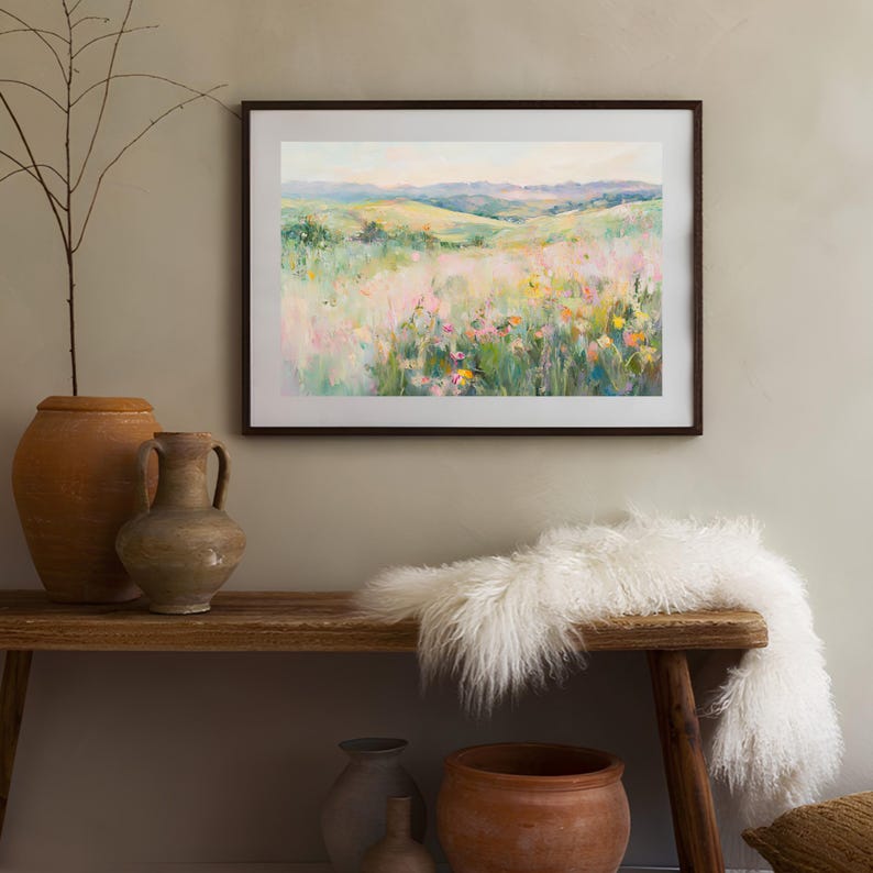 K&ouml;nnte beinhalten: Ein gerahmtes Gem&auml;lde, das ein Feld mit Wildblumen zeigt, im Hintergrund rollen die H&uuml;gel. Das Gem&auml;lde ist in einem braunen Rahmen und h&auml;ngt an einer beigefarbenen Wand. Das Gem&auml;lde ist umgeben von Einrichtungsgegenst&auml;nden, darunter eine Vase, ein Krug, ein wei&szlig;er Fell&uuml;berwurf und eine Holzbank.