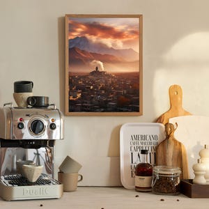 May include: A framed photograph of a mountain village with a red and orange sunset sky. The village is nestled in a valley with a mountain in the background. The photo is hanging on a wall above a coffee machine and other kitchen items.