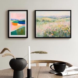 K&ouml;nnte beinhalten: Zwei gerahmte abstrakte Landschaftsbilder, eines mit einem farbenfrohen geometrischen Design und das andere mit einem weichen, impressionistischen Stil. Die Bilder h&auml;ngen an einer wei&szlig;en Wand &uuml;ber einem Holztisch mit einer schwarzen Vase, einer wei&szlig;en Kerze und einer schwarzen Kaffeetasse.