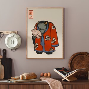 Op de afbeelding: Ingelijste kunstafdruk met een cartoonkikker in een rode kimono met witte wolkenpatronen, die een waaier vasthoudt. Japanse tekst in de linkerbovenhoek. De kikker heeft een serieuze uitdrukking. Het kunstwerk wordt in een keuken getoond.