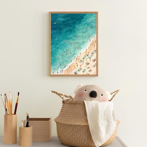 May include: A framed illustration of a beach scene with a blue ocean, white sand, and many people swimming and sunbathing. The illustration is in a wooden frame and is hanging on a white wall.