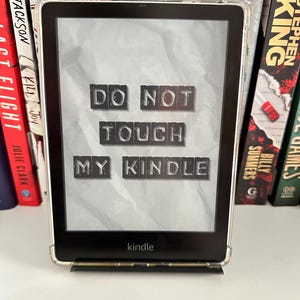 May include: A black Kindle e-reader with a clear protective case, displaying the text "DO NOT TOUCH MY KINDLE" in block letters. The e-reader is propped up on a stand, with a bookshelf in the background.