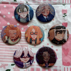 Könnte beinhalten: Sieben runde Ansteckbuttons mit farbenfrohen Illustrationen von Charakteren im Anime-Stil. Die Buttons zeigen Porträts verschiedener Charaktere mit unterschiedlichen Frisuren, Gesichtsausdrücken und Accessoires. Die Buttons sind auf einem rosa-weiß geblümten Stoff angeordnet.
