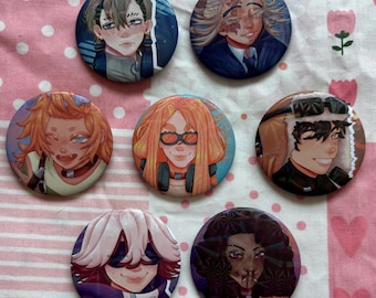 May include: Seven circular pin badges featuring anime-style character portraits. The badges showcase diverse characters with varied hairstyles, expressions, and accessories. The badges are displayed on a pink and white floral patterned fabric.