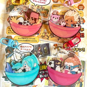 Könnte beinhalten: Vier bunte Schlüsselanhänger mit Comicfiguren in klaren Plastikkugeln. Die Figuren stammen aus der Anime-Serie Inazuma Eleven. Die Schlüsselanhänger sind an Metallringen befestigt.