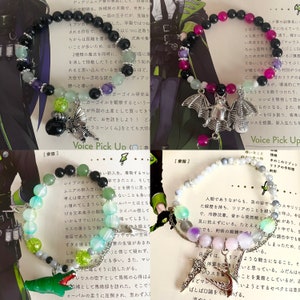 Può includere: Due braccialetti con ciondoli. Il primo braccialetto ha perline nere, verdi e rosa con un ciondolo a forma di pipistrello argentato. Il secondo braccialetto ha perline bianche, verdi e viola con un ciondolo a forma di spada argentato. Entrambi i braccialetti hanno una catena argentata attaccata al ciondolo.