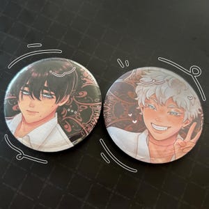 以下が含まれることがあります： アニメスタイルのイラストが描かれた丸いバッジが2つ。1つは黒髪と青い目のキャラクター、もう1つは白い髪でピースサインをしているキャラクターです。どちらも白いシャツと装飾的な背景があります。