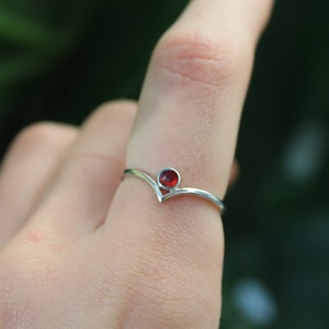 Może przedstawiać: Srebrny pierścionek z czerwonym kamieniem szlachetnym osadzonym w oprawie w kształcie litery V.