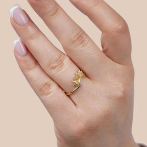 Pu&ograve; includere: Un anello dorato con un design a raggi di sole. L'anello presenta una fascia a forma di V e un motivo a raggi di sole dettagliato nella parte superiore. L'anello &egrave; mostrato su una mano, su uno sfondo rosa chiaro.