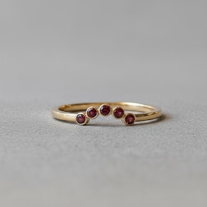 Puede incluir: Un anillo dorado con cinco pequeñas piedras preciosas redondas y rojas dispuestas en forma de arco. El anillo tiene un acabado pulido y un diseño sencillo. Las piedras están espaciadas uniformemente a lo largo del arco.