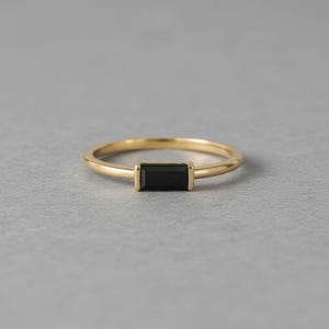 Op de afbeelding: Een gouden ring met een rechthoekige zwarte steen. De ring heeft een dunne band en een eenvoudig, elegant ontwerp. De zwarte steen is gezet in een bezel setting, getoond op een grijze ondergrond.