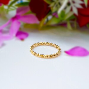 Puede incluir: Un anillo de banda de oro simple con un diseño trenzado. El anillo está sobre una superficie blanca.