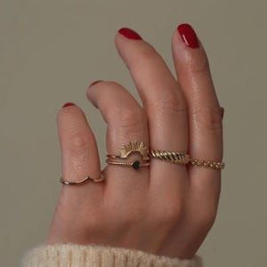 Pu&ograve; includere: Una mano con diversi anelli d'oro. Gli anelli includono una fascia ondulata, un design a raggiera con un cuore nero, uno stile a corda intrecciata e una fascia di perline. Le unghie sono dipinte di rosso.