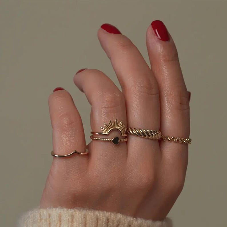 Pu&ograve; includere: Una mano con diversi anelli d'oro. Gli anelli includono una fascia ondulata, un design a raggiera con un cuore nero, uno stile a corda intrecciata e una fascia di perline. Le unghie sono dipinte di rosso.