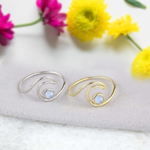 May include: Two wave-shaped rings, one silver and one gold, each featuring a small, round, light-colored gemstone. The rings are set against a soft gray background, with blurred pink and yellow flowers in the background.